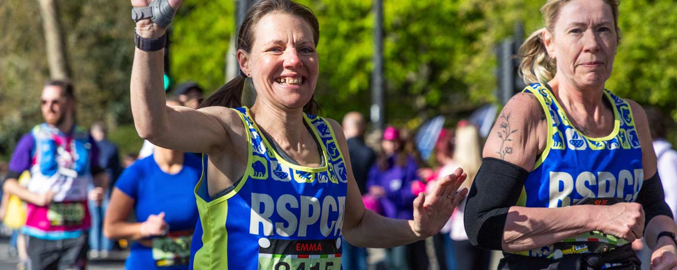 Two female charity runners wearing RSPCA branded vests waving to camera.