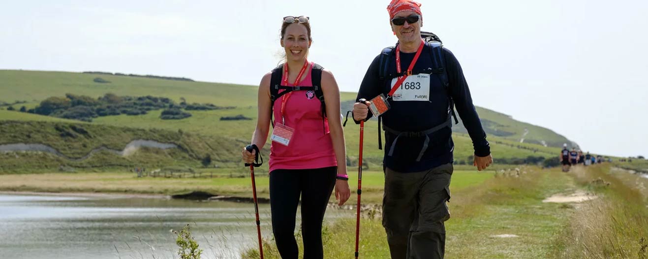 Two smiling threkkers with walking poles walking along side a lake in the Lake District.