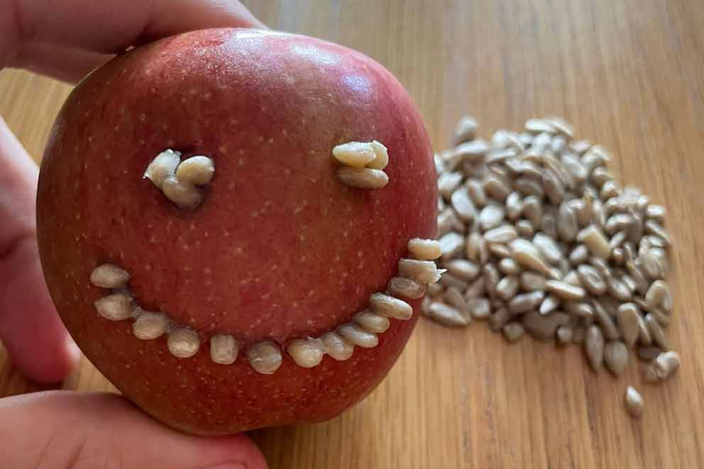 Red apple decorated with sunflower seeds arranged in a smiley face, with a pile of loose seeds on a wooden surface in the background.