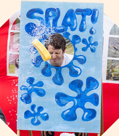 A person underneath a sign saying 'splat', their face is scrunched up as a wet sponge comes towards them.