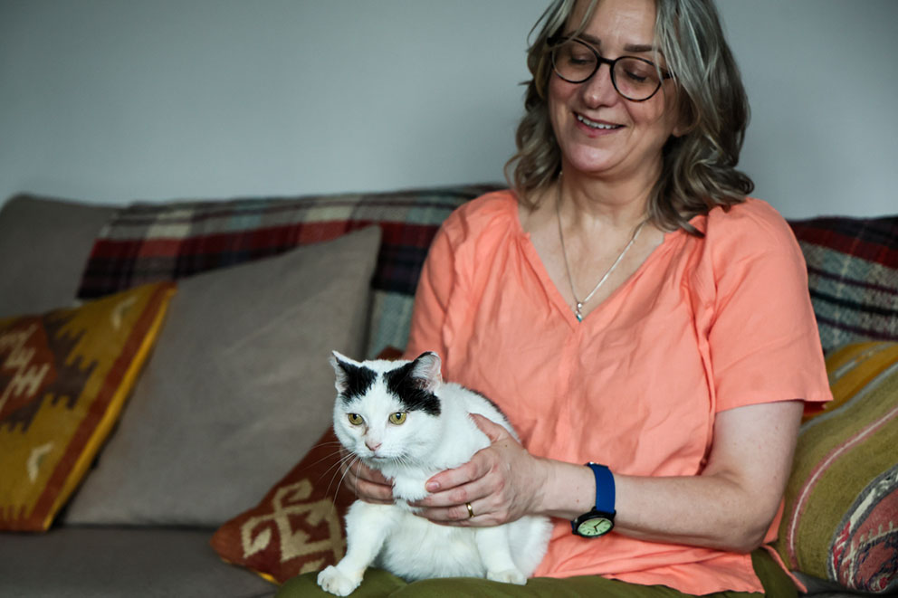 A smiling woman sits on a sofa and gently holds Molly, a black and white cat, on her lap.
