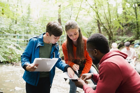 A man and two children in the woods, holding a clip board and pointing at a sheet of paper. 