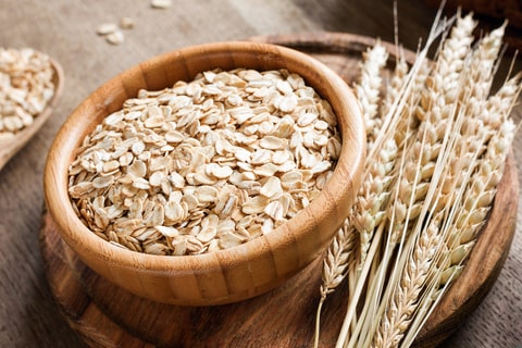 A bowl of oats in a wooden bowl, alongside a bunch of wheat. 