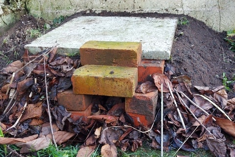 Multiple orange bricks on the ground, with a concrete slab on top, surrounded by soil.
