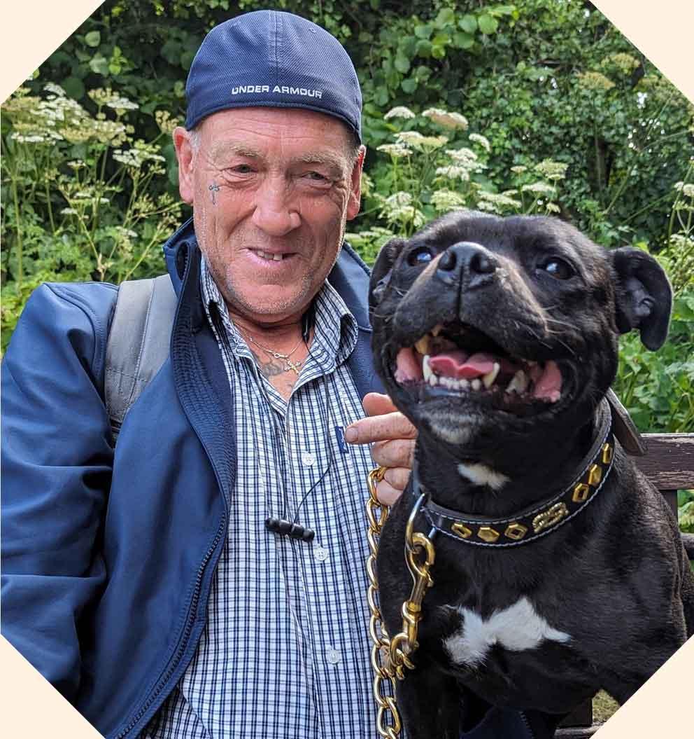 A man wearing a hat with a black dog sitting on a bench outside.