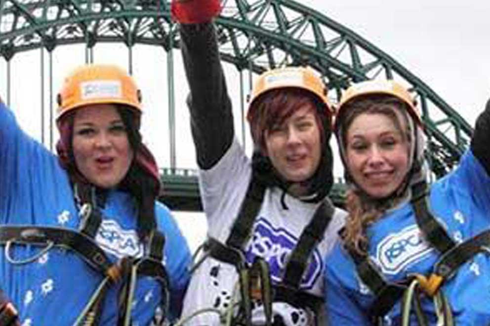 Group of fundraisers raising their arms on completing an abseiling challenge with a bridge in the background.